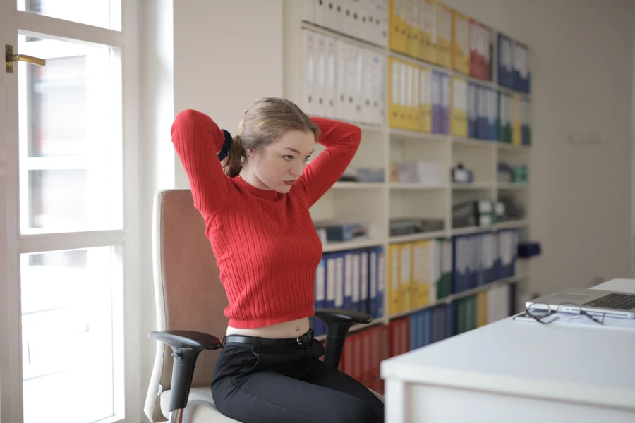 Jeune femme assise sur une chaise de bureau, mains derrière la nuque, qui ouvre la poitrine entre deux tâches