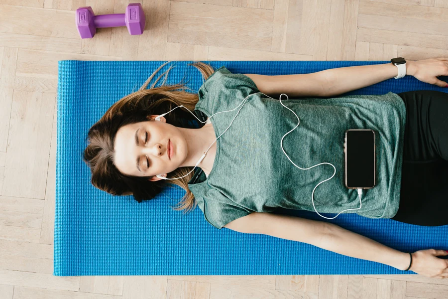 Femme allongée sur le dos sur un tapis de yoga, yeux fermés, écouteurs aux oreilles, qui suit une séance guidée